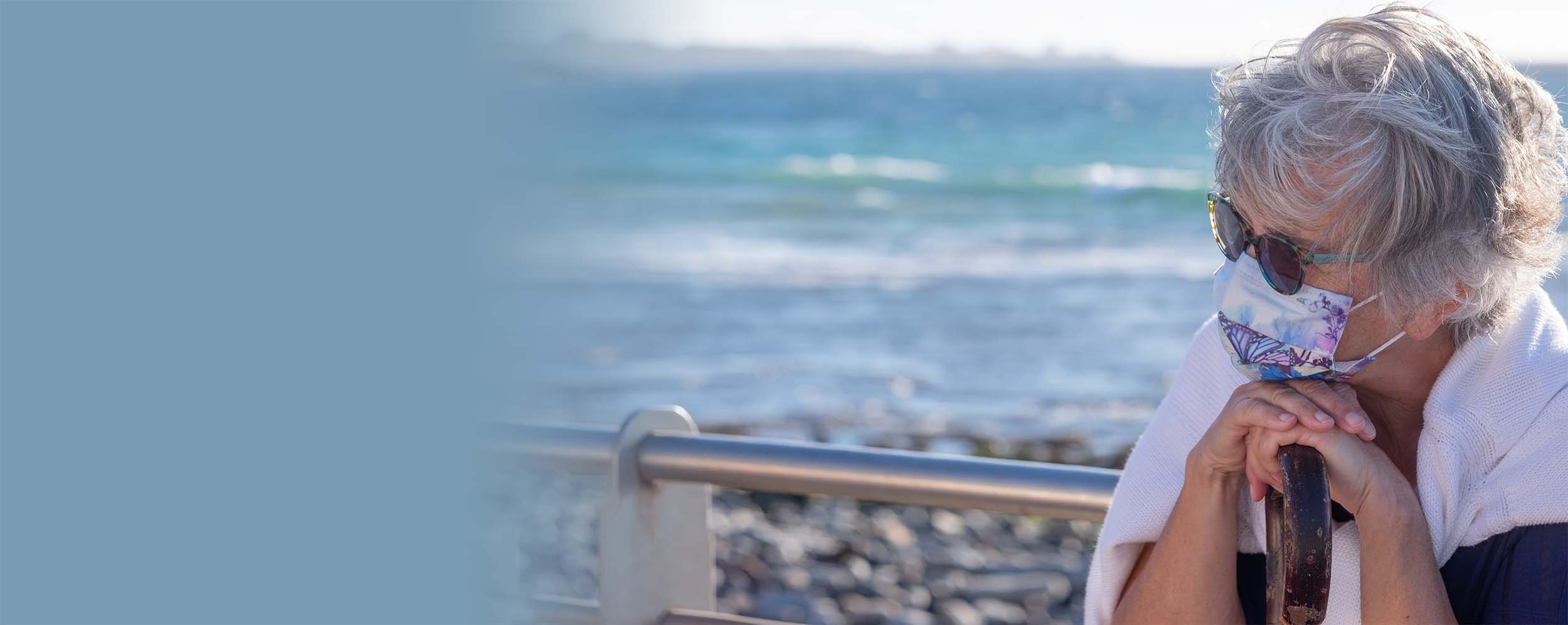 An older woman wearing a face mask and sunglasses sits by the beach, looking wistful