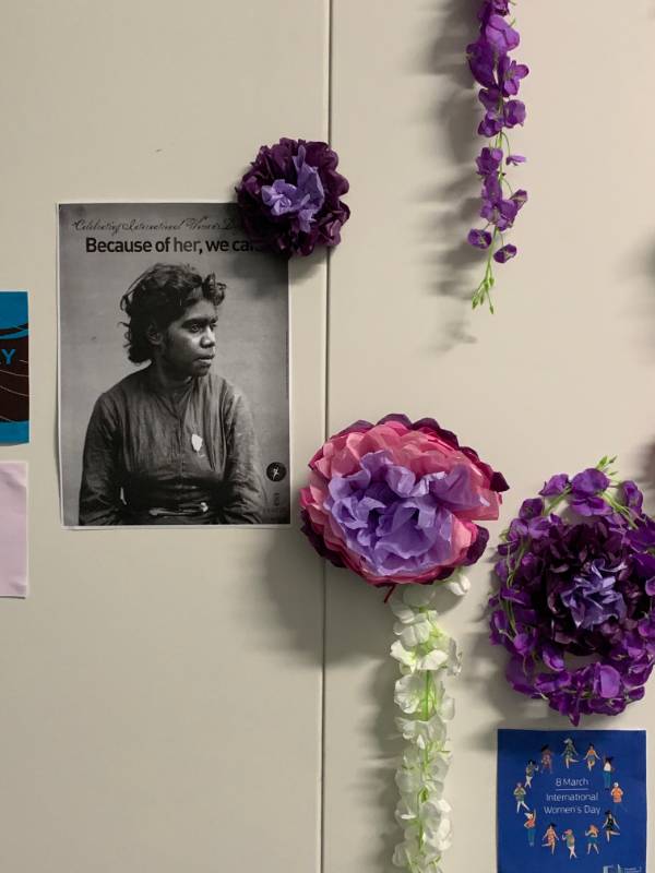 A poster with an Aboriginal woman, Because of Her We Can, and purple flowers for International Women's Day